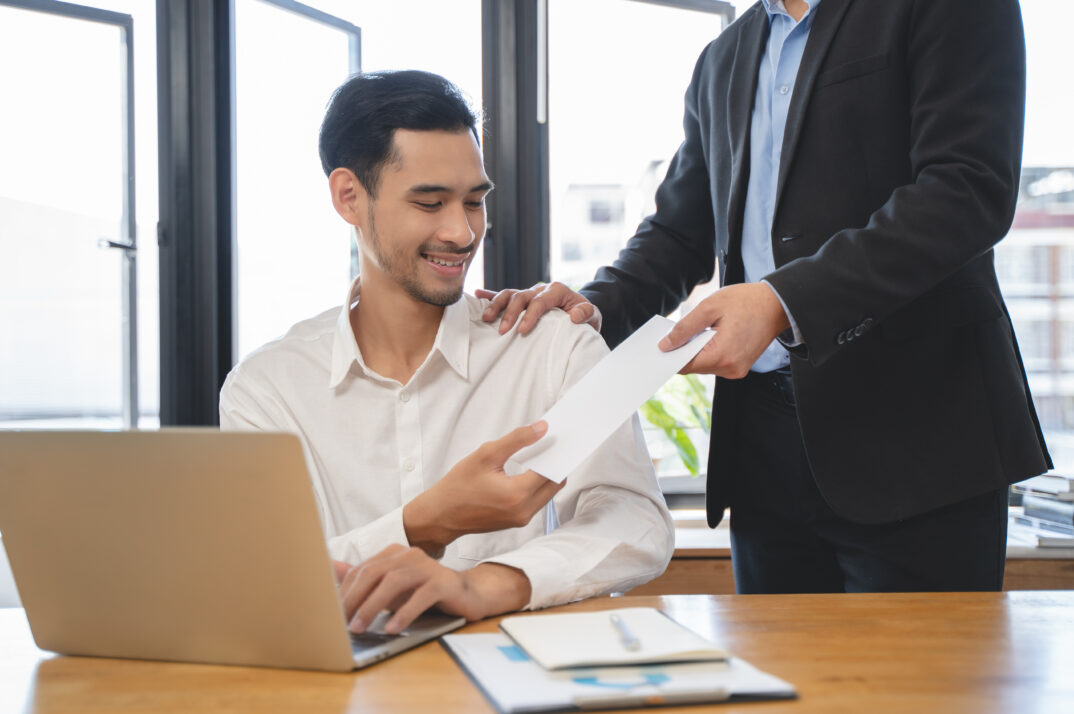 A manager hands a bonus check envelope to an employee sitting at a laptop.